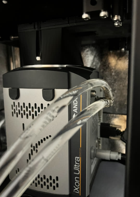 Close-up of a silver Andor iXon Ultra EMCCD camera mounted under a microscope, with clear cooling tubes running to it.