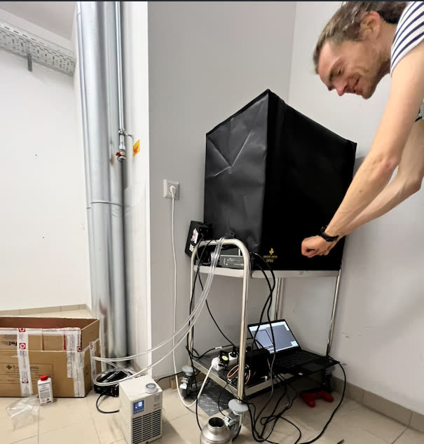 A researcher leaning over a black light-proofed enclosure standing on a trolley in a lab, with a laptop, tubing, and a chiller on the shelf beneath it.