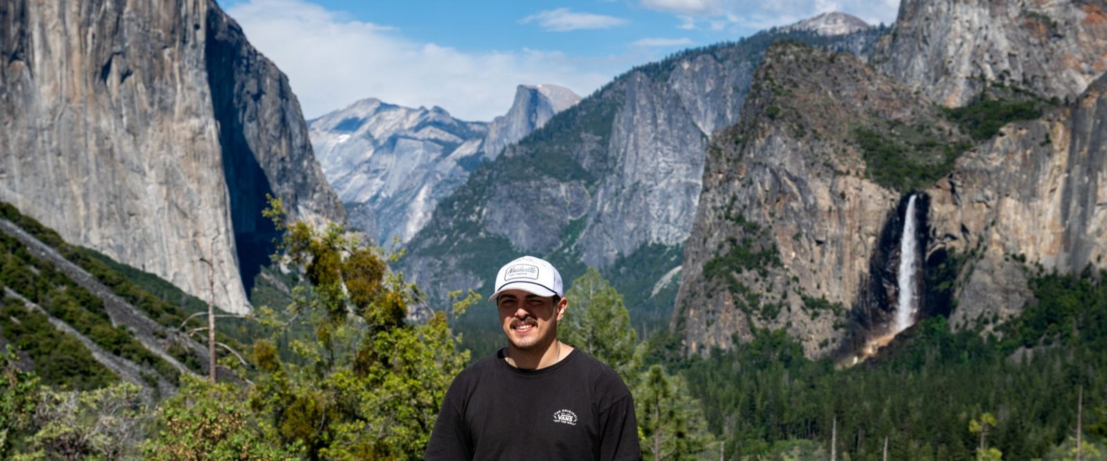 Michael Saldivar, in a white cap and a black T-shirt, stands in front of Yosemite Valley, with El Capitan on the left, Half Dome in the distance and Bridalveil Fall on the right.