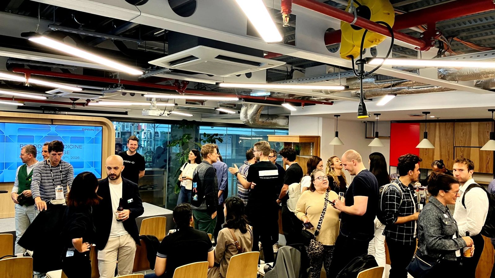 Participants standing and talking in groups in a Station F event space, a screen behind them showing the SPRIND Recoding Medicine Challenge title slide.