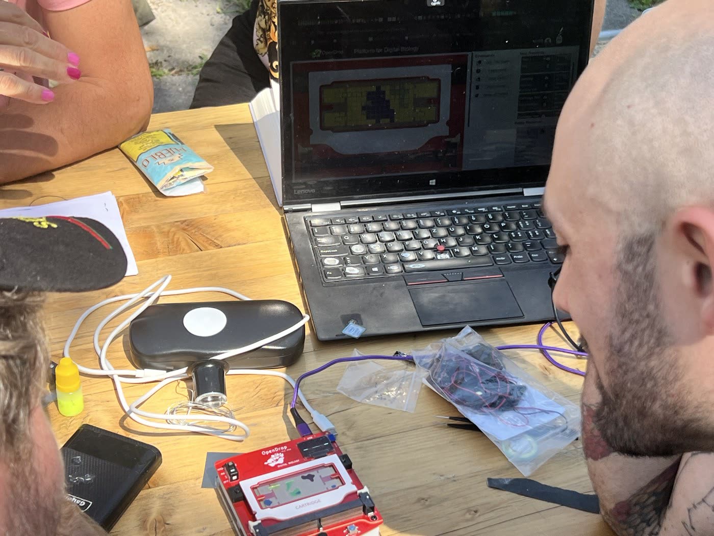 Two participants watch a laptop running the OpenDrop digital-biology interface, with the red OpenDrop cartridge board on the table.